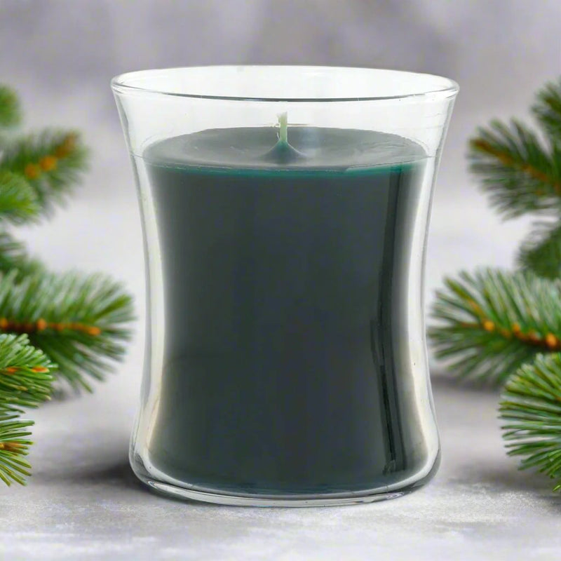 Green Christmas candle in a clear glass container with Christmas tree branches in the background