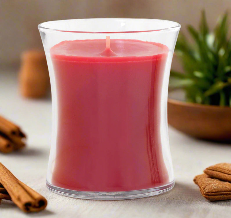 red candle in a clear elegant glass container on a surface with cinnamon sticks and a plant in the background.