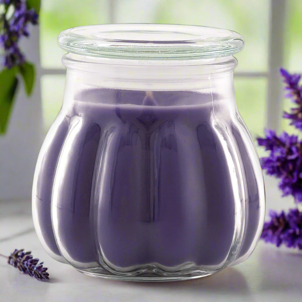purple candle contained within a clear glass jar, with a greenery background suggesting an indoor setting.