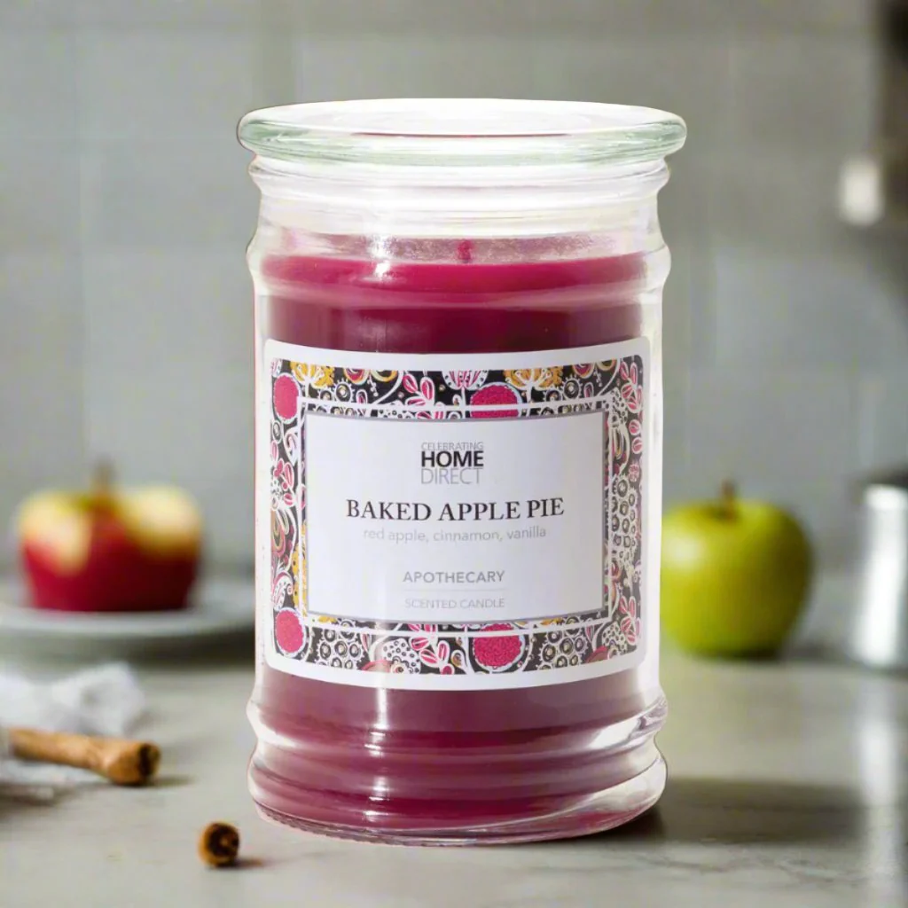 A Apothecary candle with a red hue, labeled Baked Apple Pie, placed on a kitchen countertop with apples and cinnamon sticks in the background.
