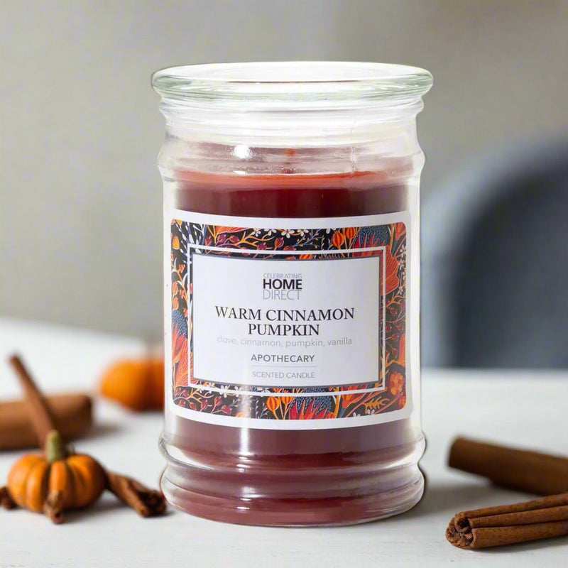 A jar candle with a label reading 'HOME WARM CINNAMON PUMPKIN APOTHECARY', surrounded by decorative pumpkins and cinnamon sticks.
