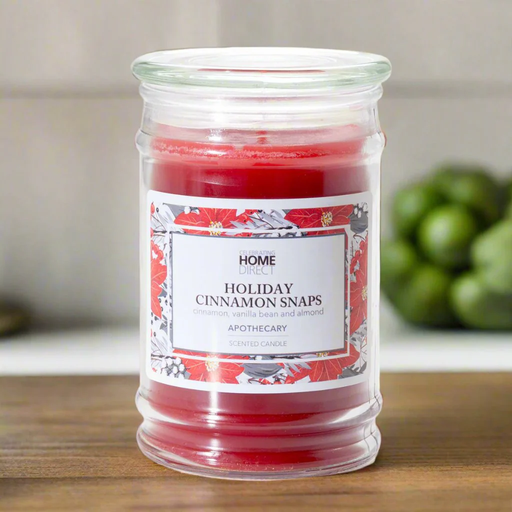 A red Apothecary candle with a label that reads 'HOME HOLIDAY CINNAMONSNAPS'. The candle is placed on a countertop with some greenery in the background.