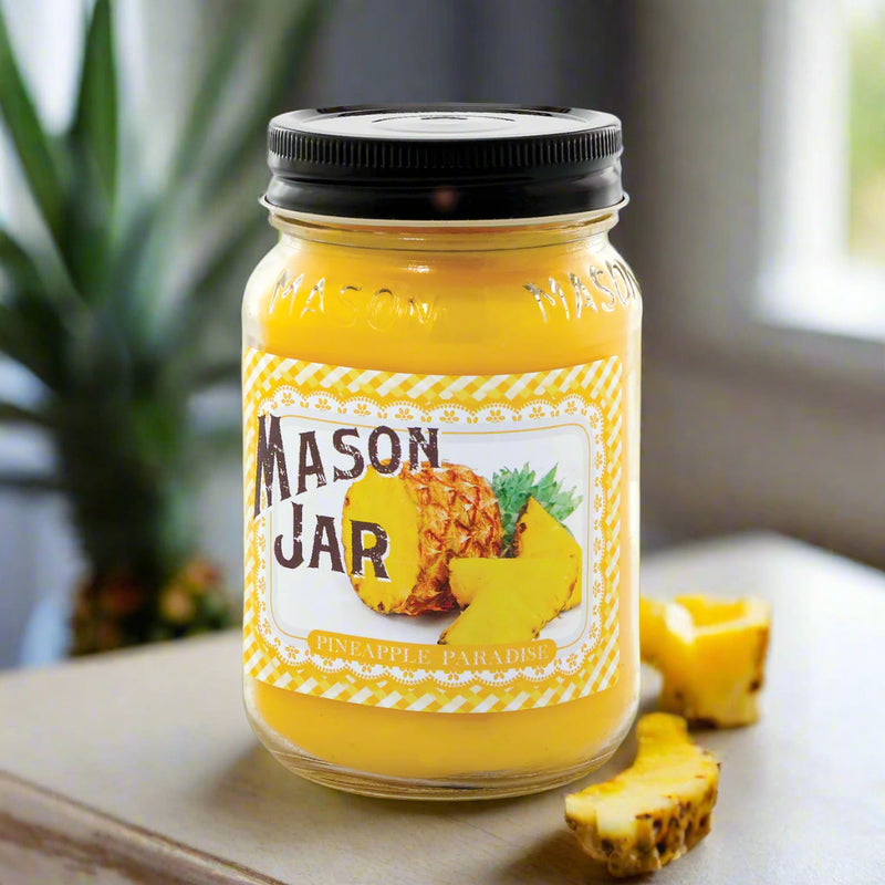 A Mason Jar Candle with a yellow label featuring a pineapple illustration, sitting on a wooden surface with a pineapple fruit in the background.