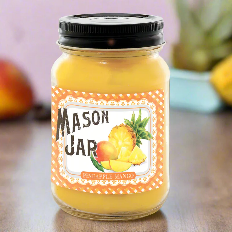 A Mason jar candle with a yellow hue and a label that reads 'Mason Jar Pineapple Mango'. There is a pineapple and mango illustration on the label.