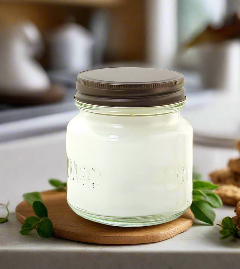 A white, 8 oz. Mason jar candle with a metal lid, filled with butter cookie scented wax, on a wooden surface with mint leaves scattered around.