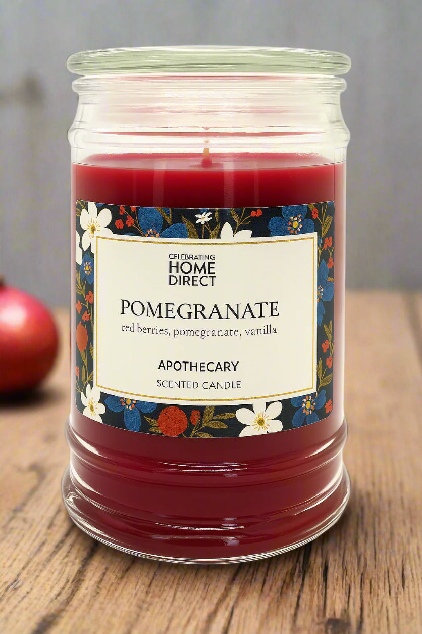 A pomegranate-scented Apothecary candle with red and white label, placed on a wooden surface with apples in the background.