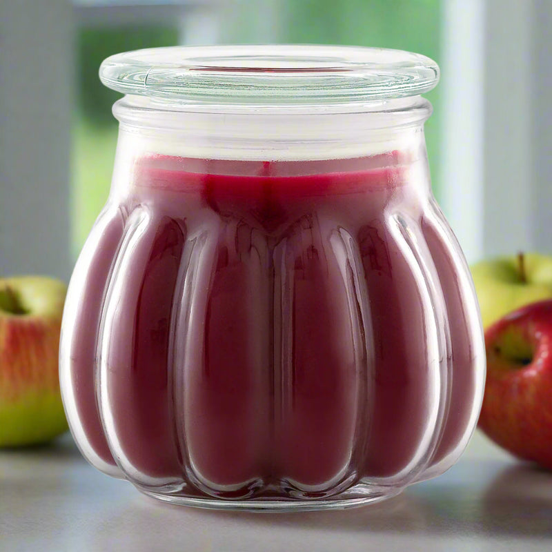A Contempo jar candle with a red hue, resembling the color of baked apple pie, placed on a surface with apples in the background.