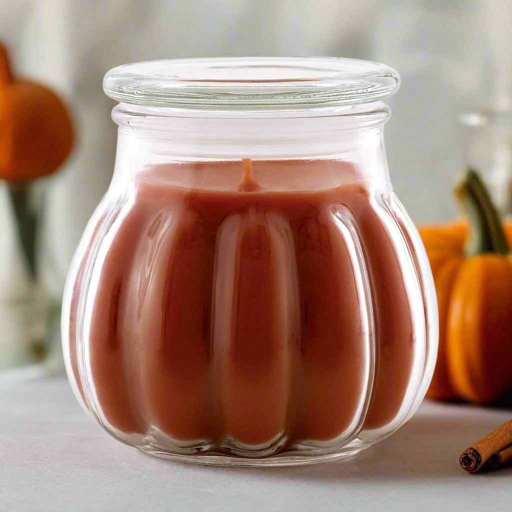 A Contempo Candle with a warm cinnamon pumpkin scent, shown in a clear glass jar with a brownish-red colored candle inside, set on a table with pumpkins and cinnamon sticks in the background.
