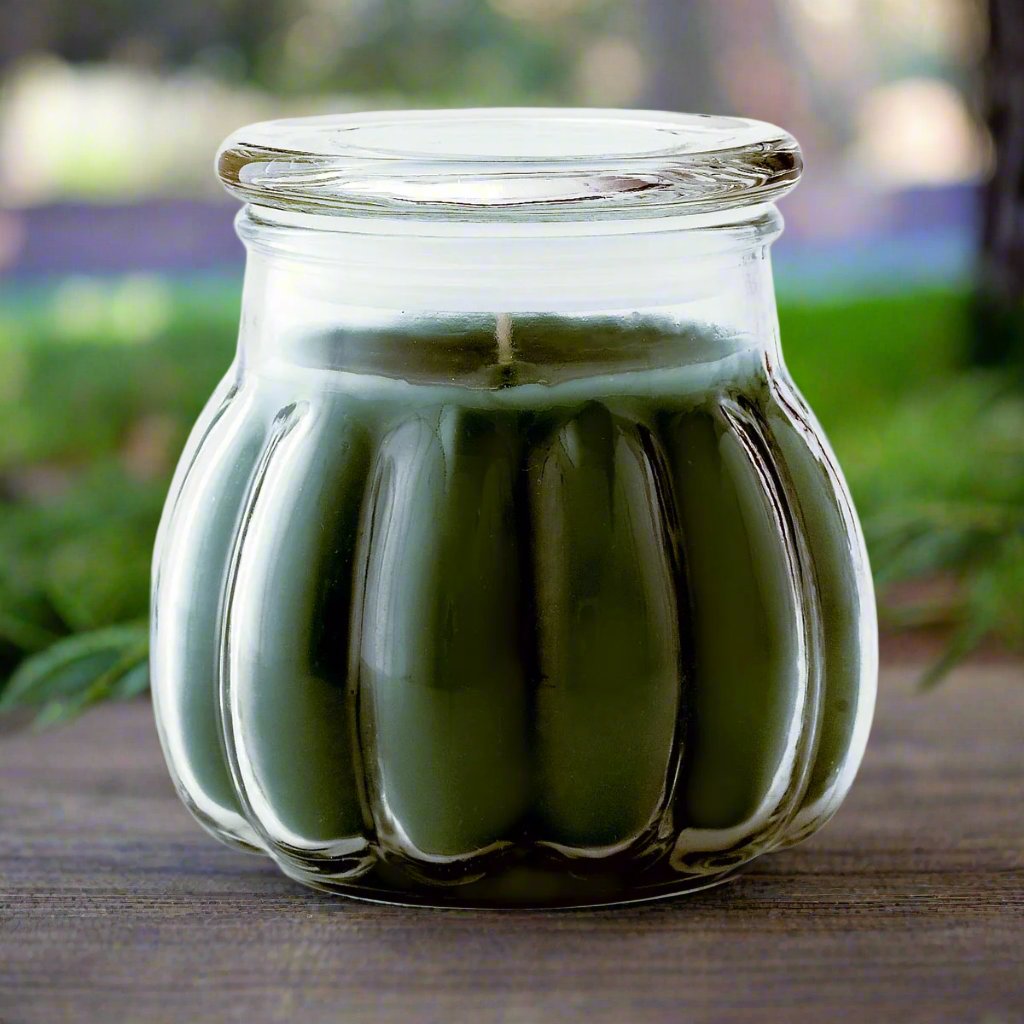 A green tinted ice evergreen Christmas candle in a clear glass jar, set outdoors with foliage in the background.