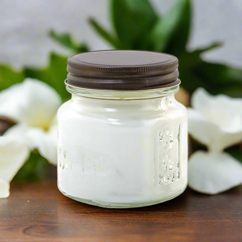 A white candle in a clear glass jar with a brown lid, placed on a wooden surface with green leaves and flowers in the background.