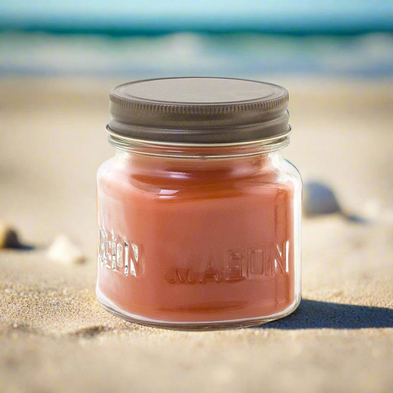 A clear glass jar candle with a brown lid, labeled 'MASON', filled with red-colored wax, displayed on a sandy beach with ocean waves in the background.