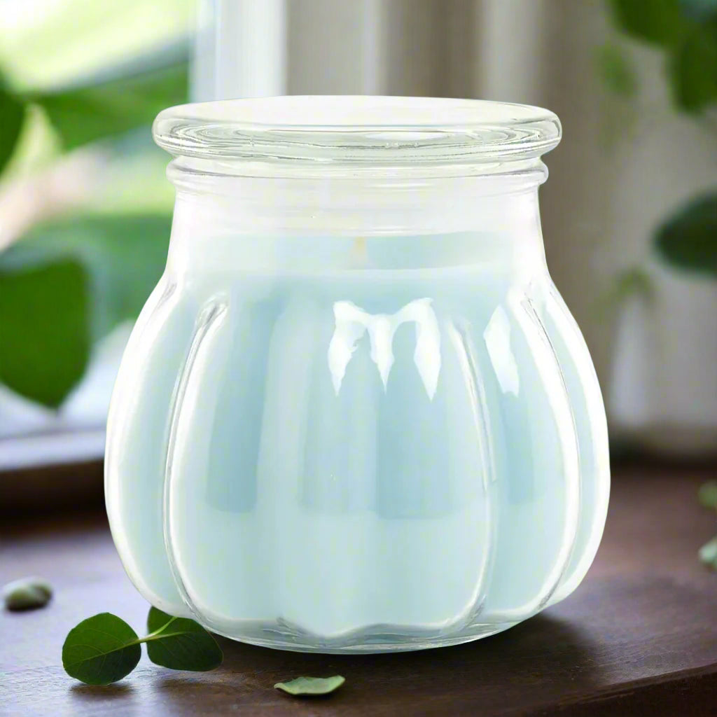 clear glass jar Contempo candle with a light sky blue tinted wax inside, set on a wooden surface with eucalyptus leaves scattered around, with a blurred background.