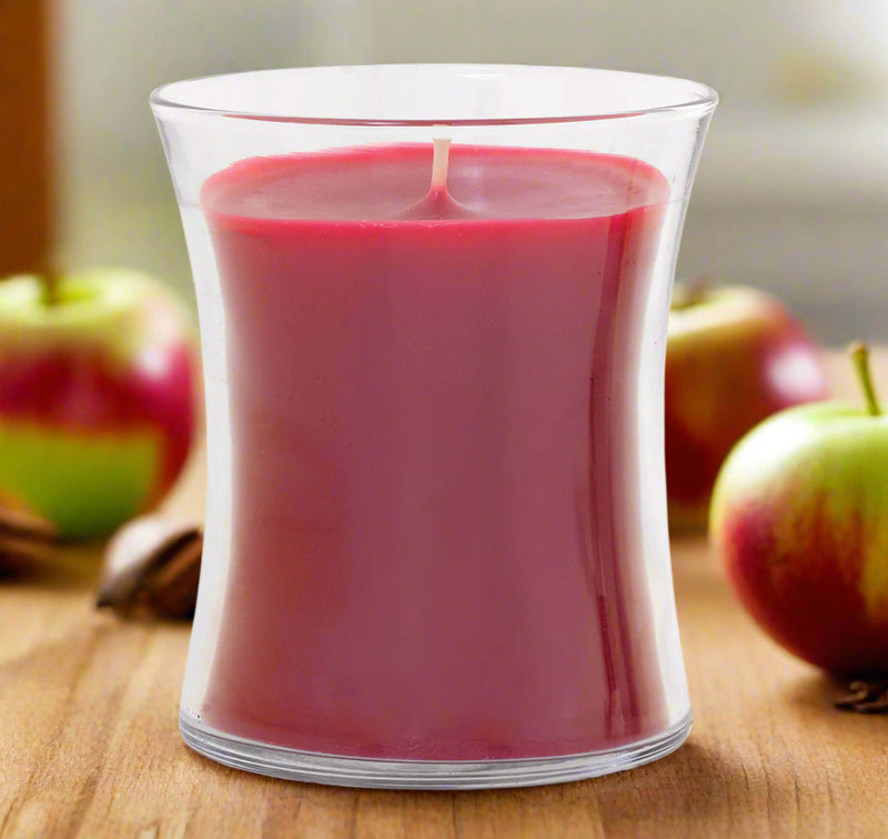 red candle in a clear glass container with apples in the background on a wooden surface