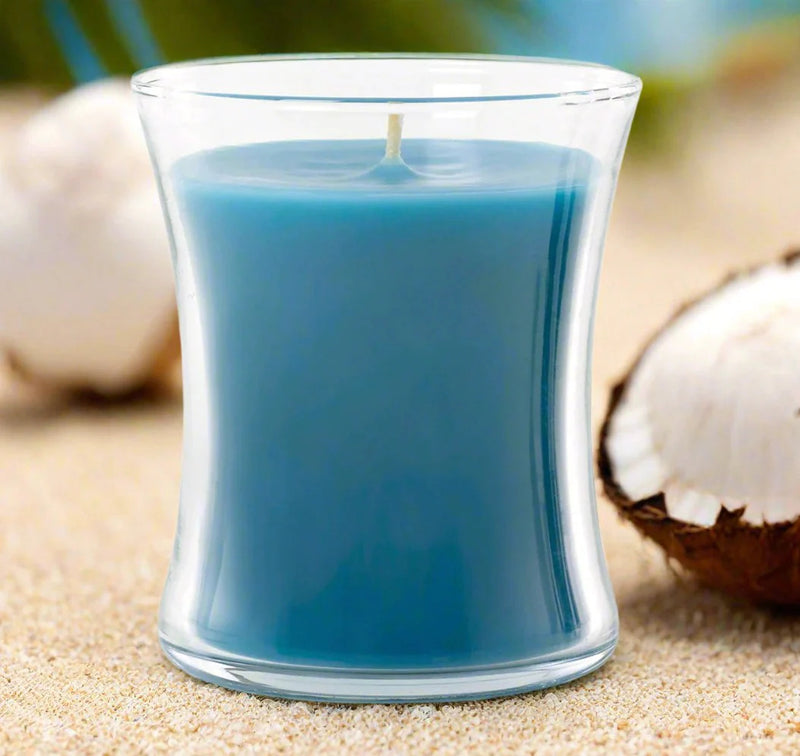 Blue elegant candle in a clear glass container on sand with coconuts in the background