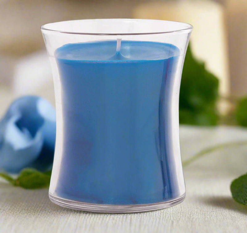 A clear glass candle with a blue wax layer, placed on a table with a blue flower and green leaves in the background.