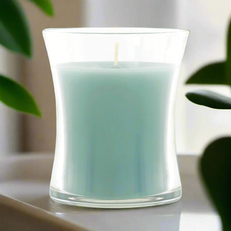 Light blue candle in elegant glass on a white surface with green plants in the background