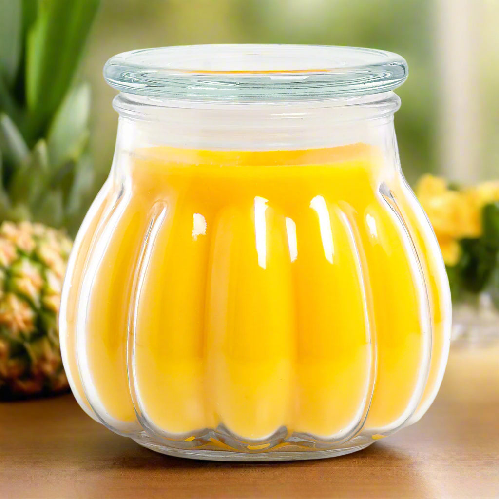 A clear glass jar candle with a yellow-hued wax inside, set on a wooden surface with pineapples and orchids in the background.